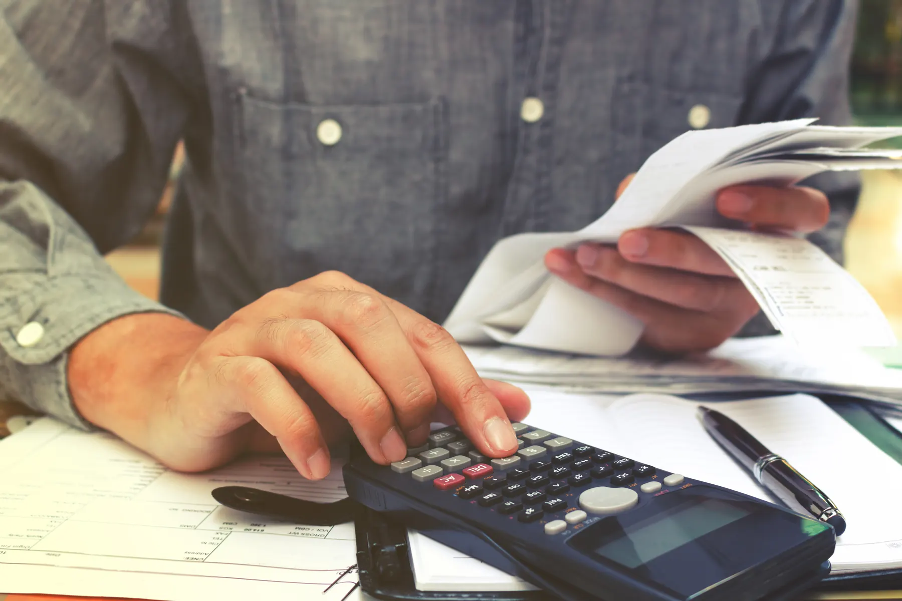 Hands working a desk calculator beside a fan of receipts, a ruled ledger and a pen