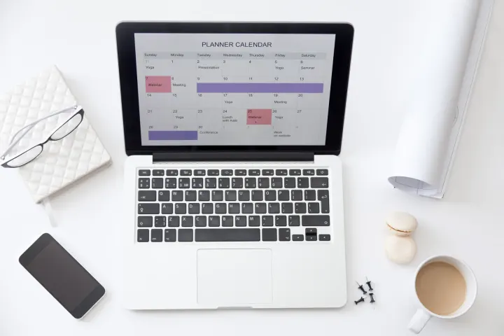 A planner calendar open on a laptop, with a notebook, glasses and coffee