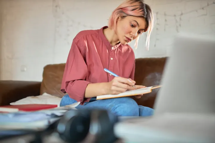 Someone writing in a notebook on a sofa, laptop beside them
