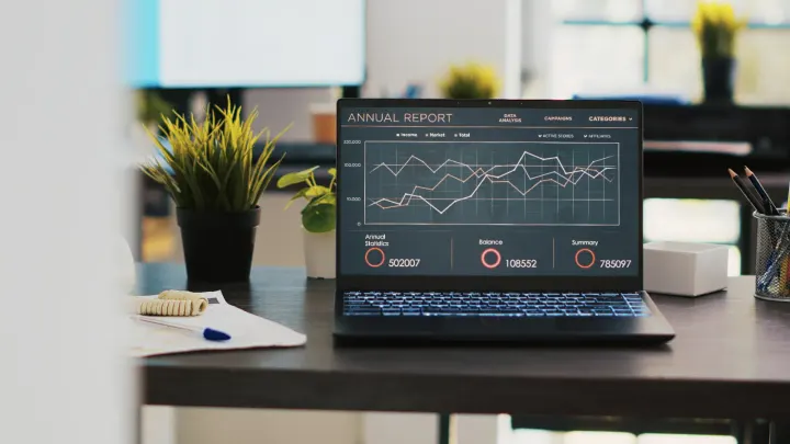 A laptop on an office desk showing an annual report dashboard of line charts and figures
