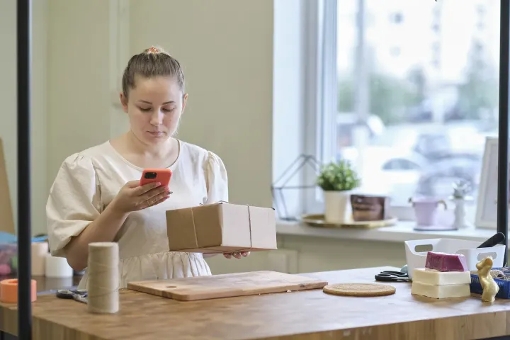 A seller checking her phone while packing small boxes at a desk