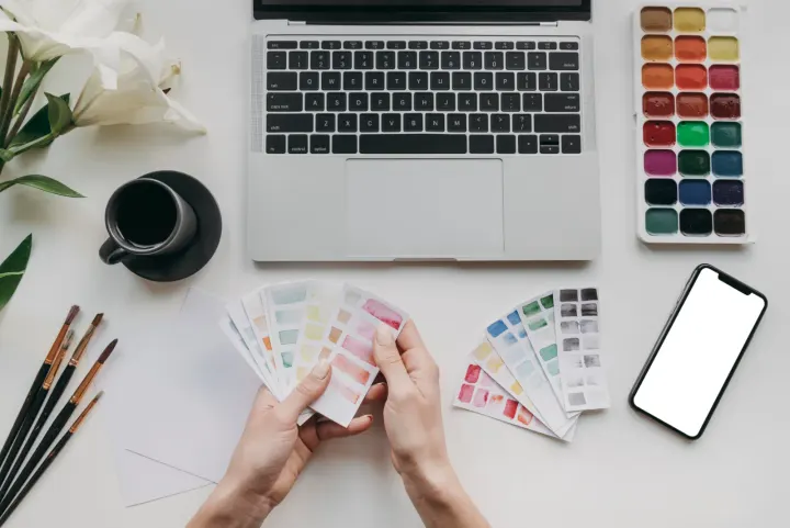 Hands holding fanned-out colour swatch cards on a desk beside a laptop and paint sets