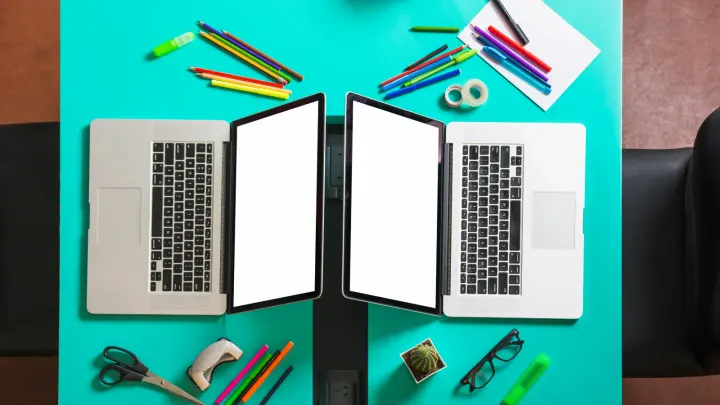 Two near-identical open laptops facing each other on a turquoise desk