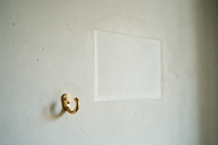 An empty brass picture hook on a pale wall, with the faded rectangle where a framed print used to hang