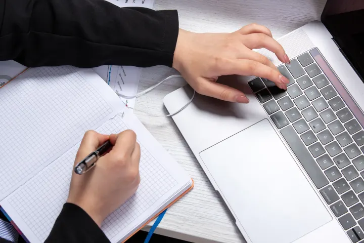 Writing notes by hand beside an open laptop